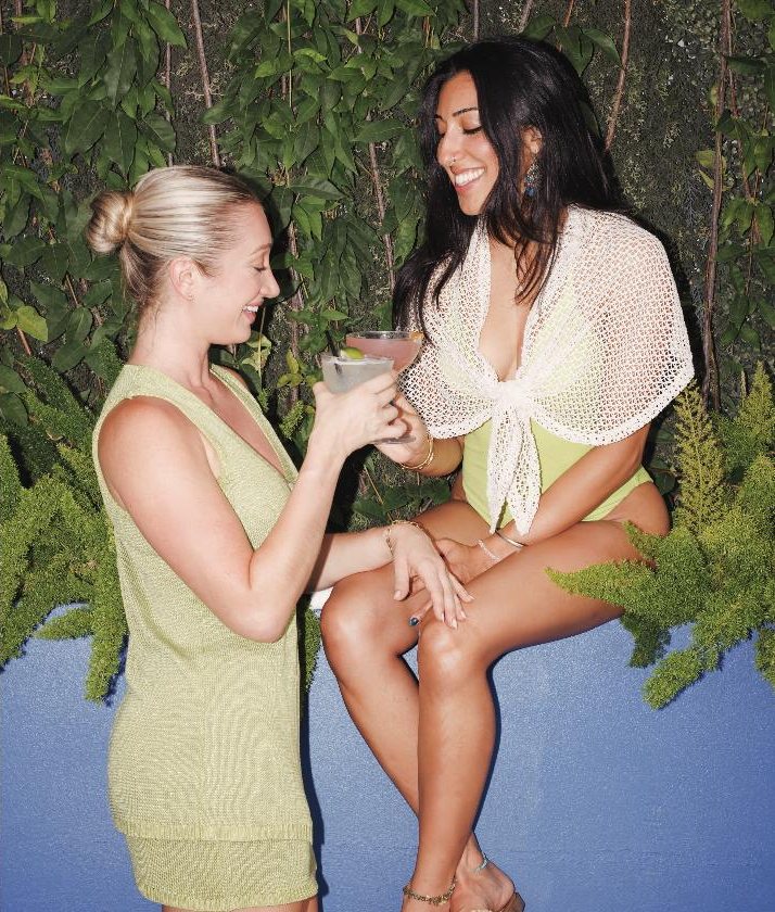 Two friends with cocktails in front of a green wall